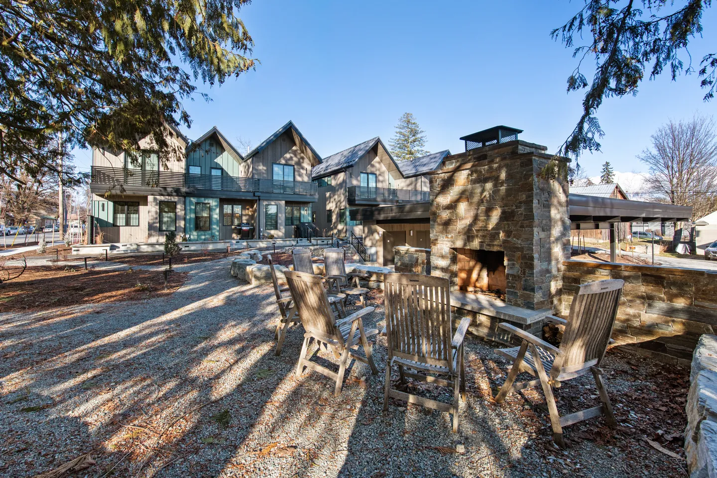 Outdoor gravel patio featuring a stone fireplace and wooden chairs in front of a modern multi-story residential building.