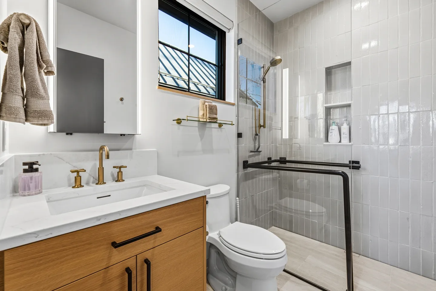 Modern bathroom featuring a light wood vanity, white marble countertop with gold faucet, toilet, and glass walk-in shower with vertical tile.