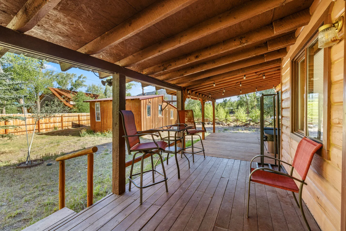 Covered wooden porch with rustic wood railings, exposed rafters, and outdoor seating overlooking a yard and outbuilding.