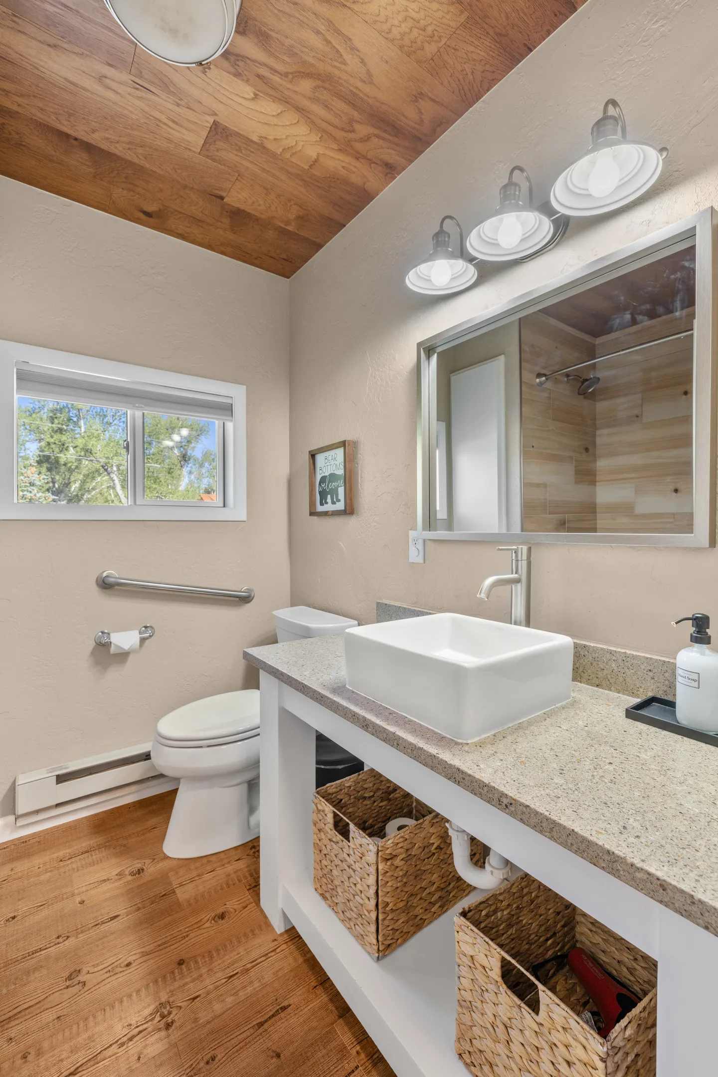 Bathroom with a vessel sink vanity, mirror, toilet with grab bar, and wood-paneled ceiling.