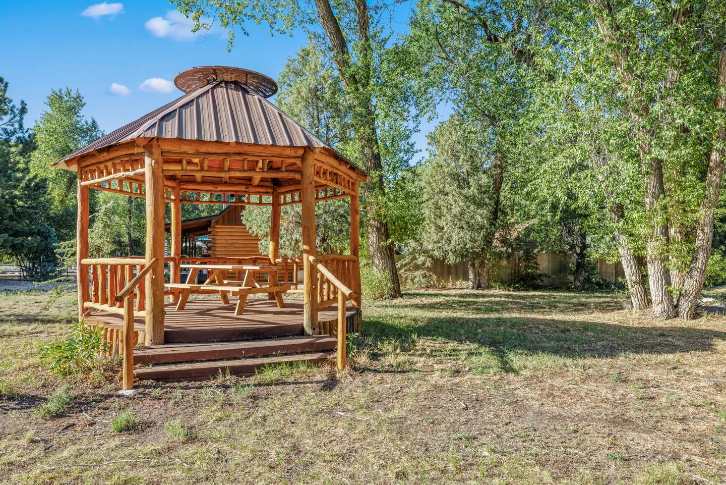 A rustic wooden gazebo with a picnic table inside, set in a grassy yard surrounded by trees under a clear blue sky.