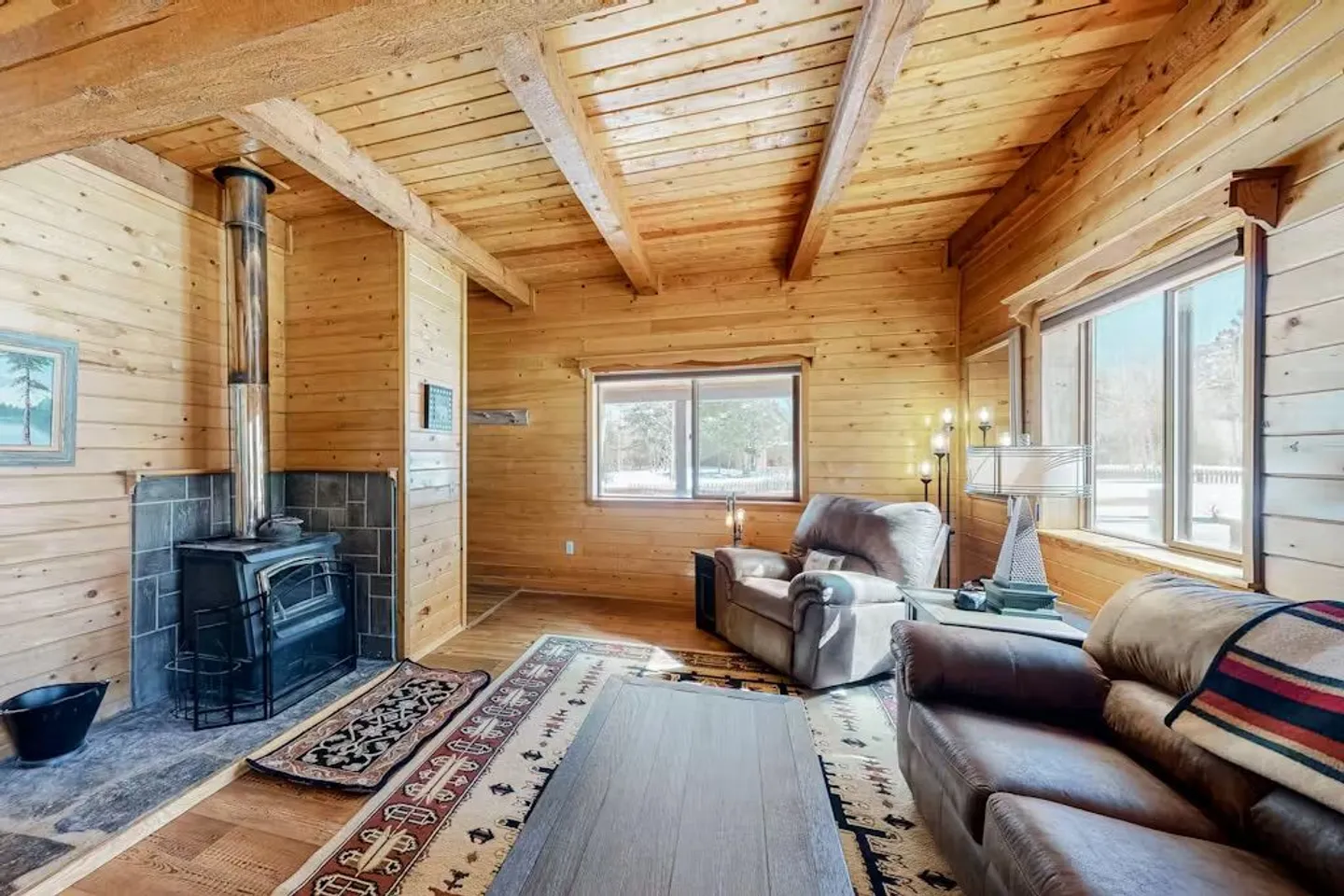 Living room with wood-paneled walls and ceiling, a wood-burning stove, patterned area rugs, a recliner, and a sofa.