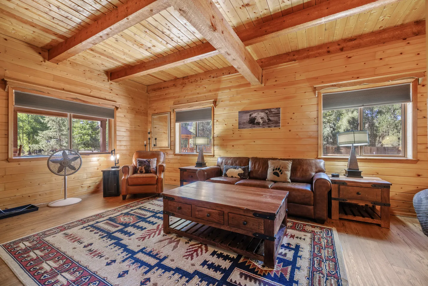 Rustic living room with wood-paneled walls, exposed ceiling beams, a leather sofa, armchair, and wooden coffee table on a patterned area rug.