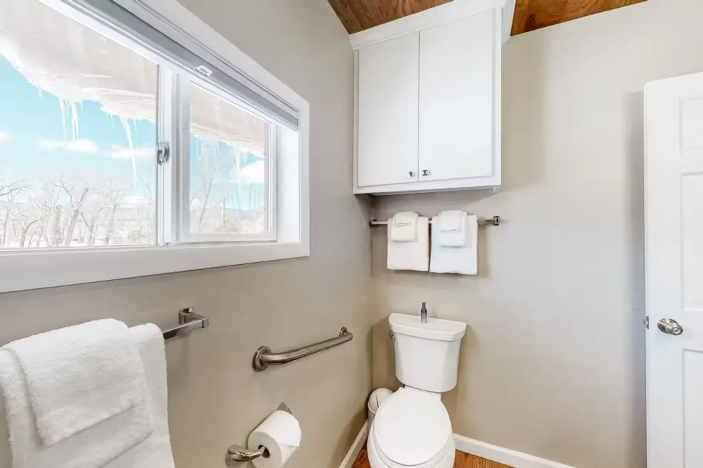 Bathroom with a toilet, safety grab bar, towel racks, upper white cabinet, and a window with icicles visible outside.
