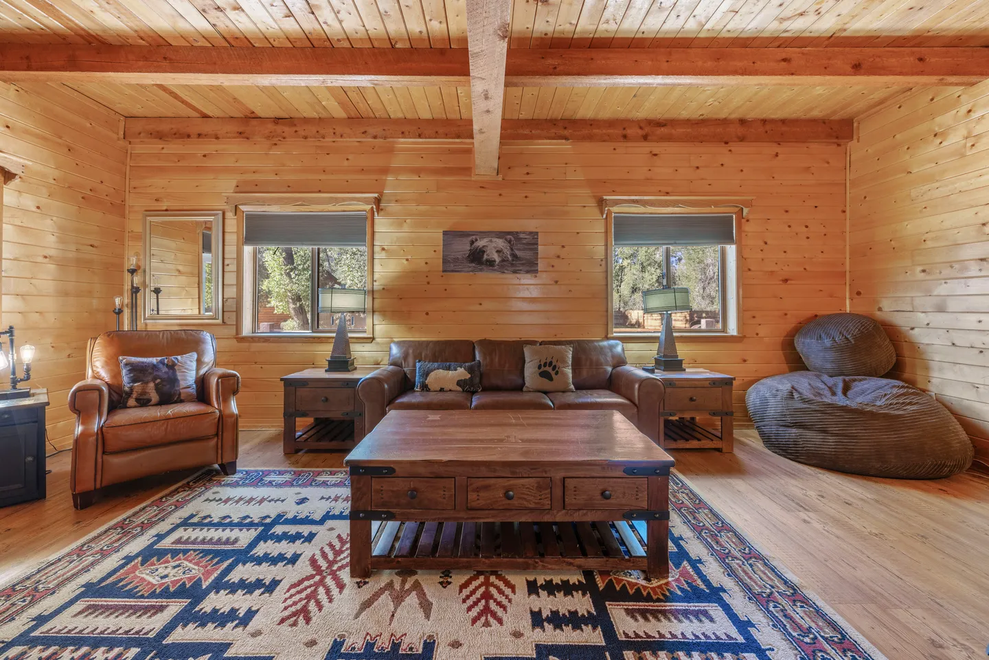 Living room with wood-paneled log walls, wood-beam ceiling, leather couch, leather armchair, large coffee table, and patterned rug.