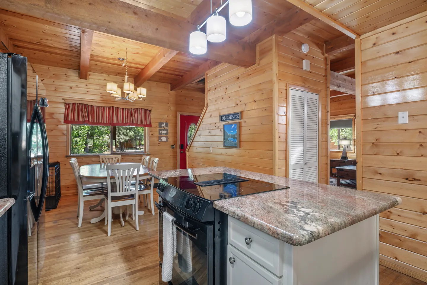 Open-concept cabin interior looking past a kitchen island with a stovetop toward a wooden dining table and chairs, wood-paneled walls, and exposed ceiling beams.