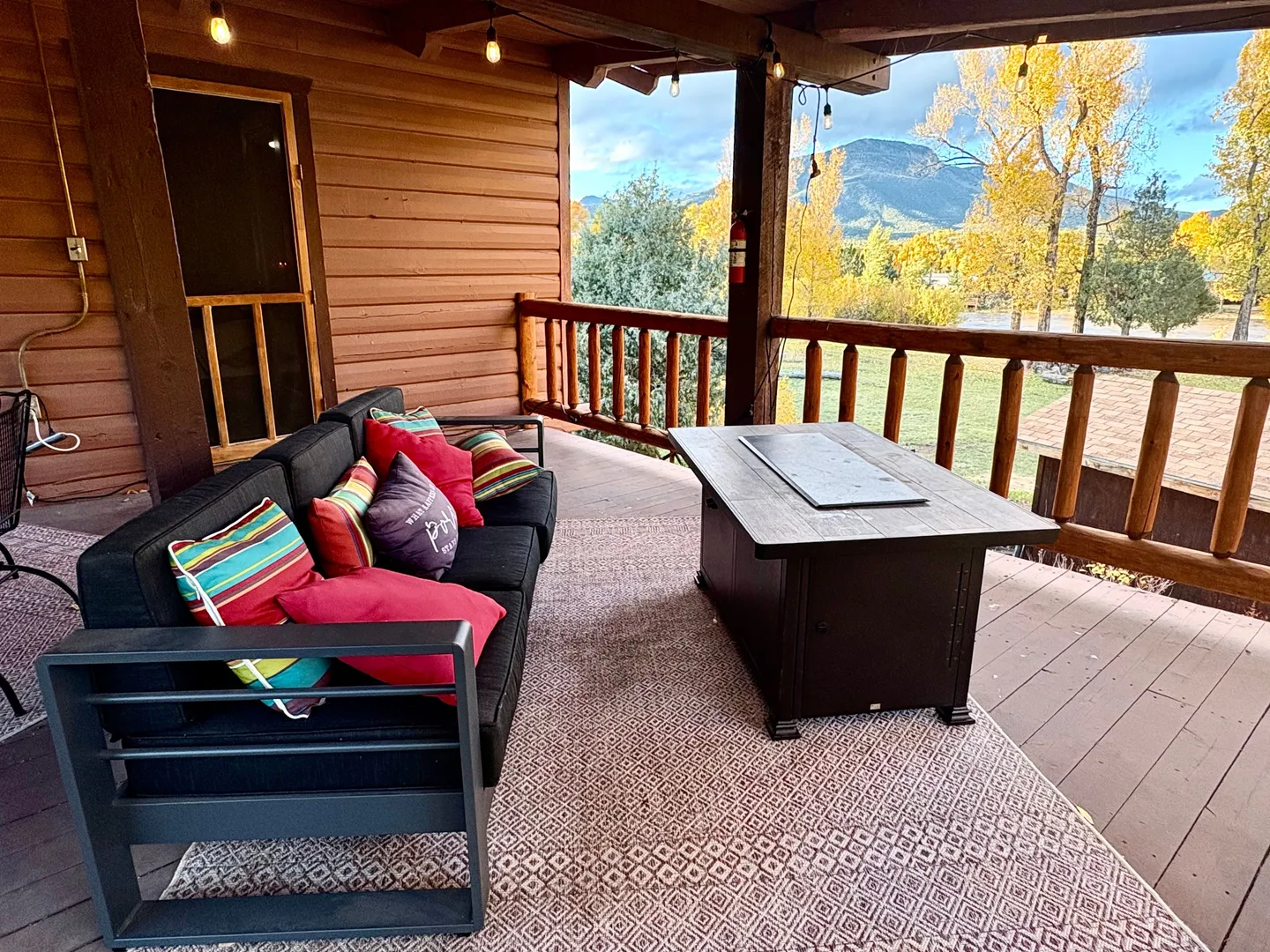 Covered outdoor porch with a cushioned sofa, fire table, patterned rug, and wooden log railing overlooking mountains and trees.