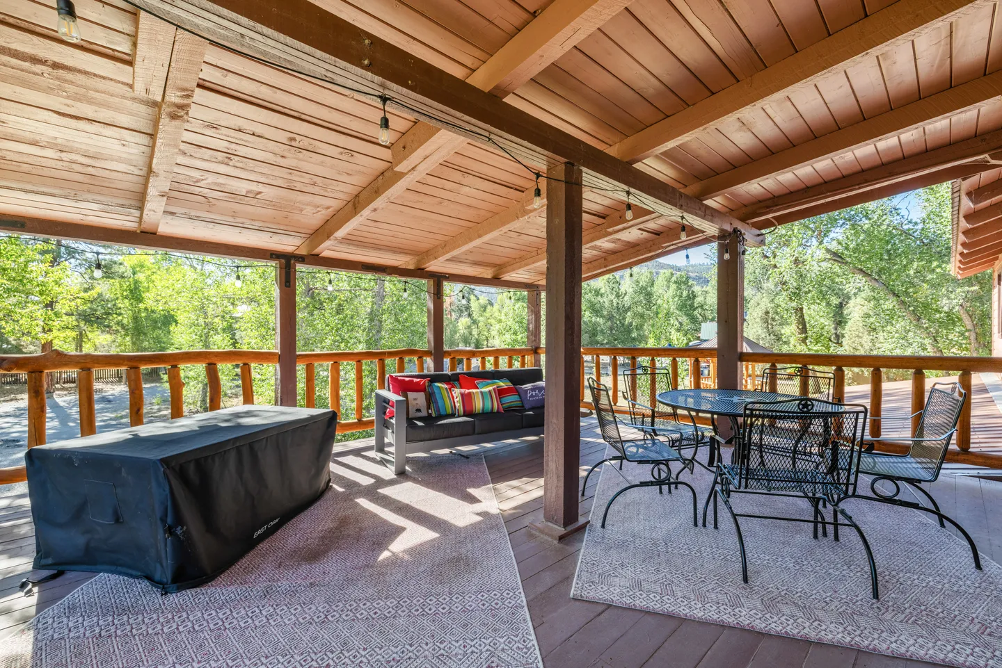 Covered outdoor deck with exposed wood rafters, outdoor dining table with chairs, an outdoor couch, and a view of surrounding trees.