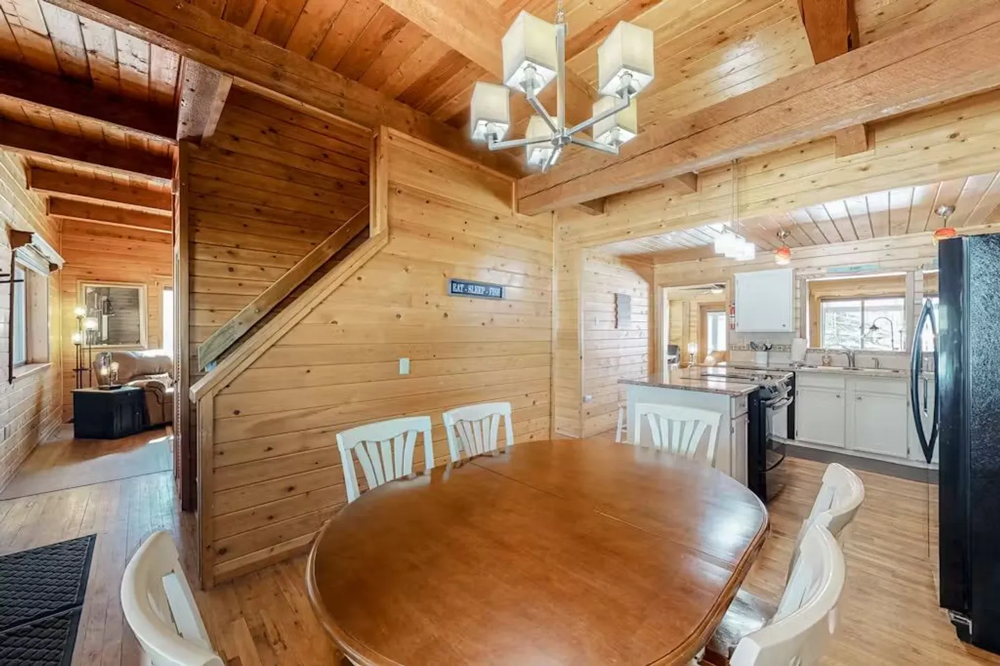 Dining area with a wooden oval table and white chairs in a wood-paneled cabin with an adjacent kitchen and staircase.