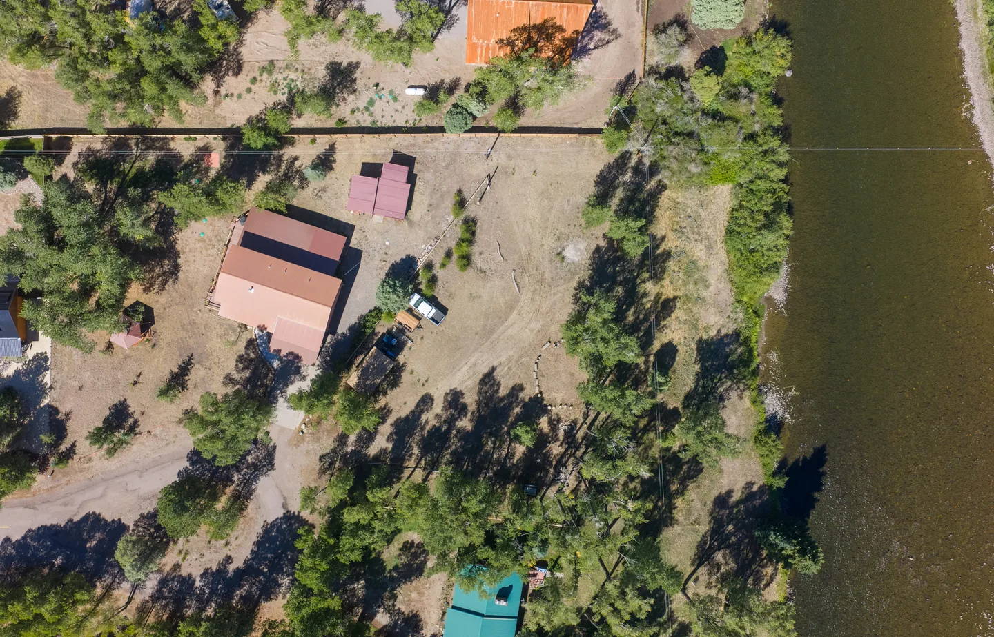 Aerial overhead view of a home with a red roof on a riverfront lot with trees and parked vehicles.