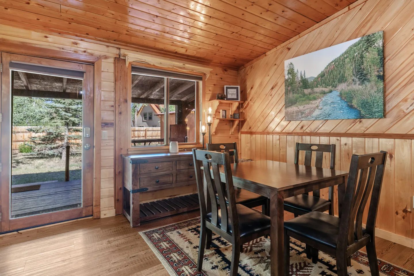 Dining area with wood-paneled walls and ceiling, featuring a dark wood dining table, matching chairs, a console table under a window, and an exterior door.