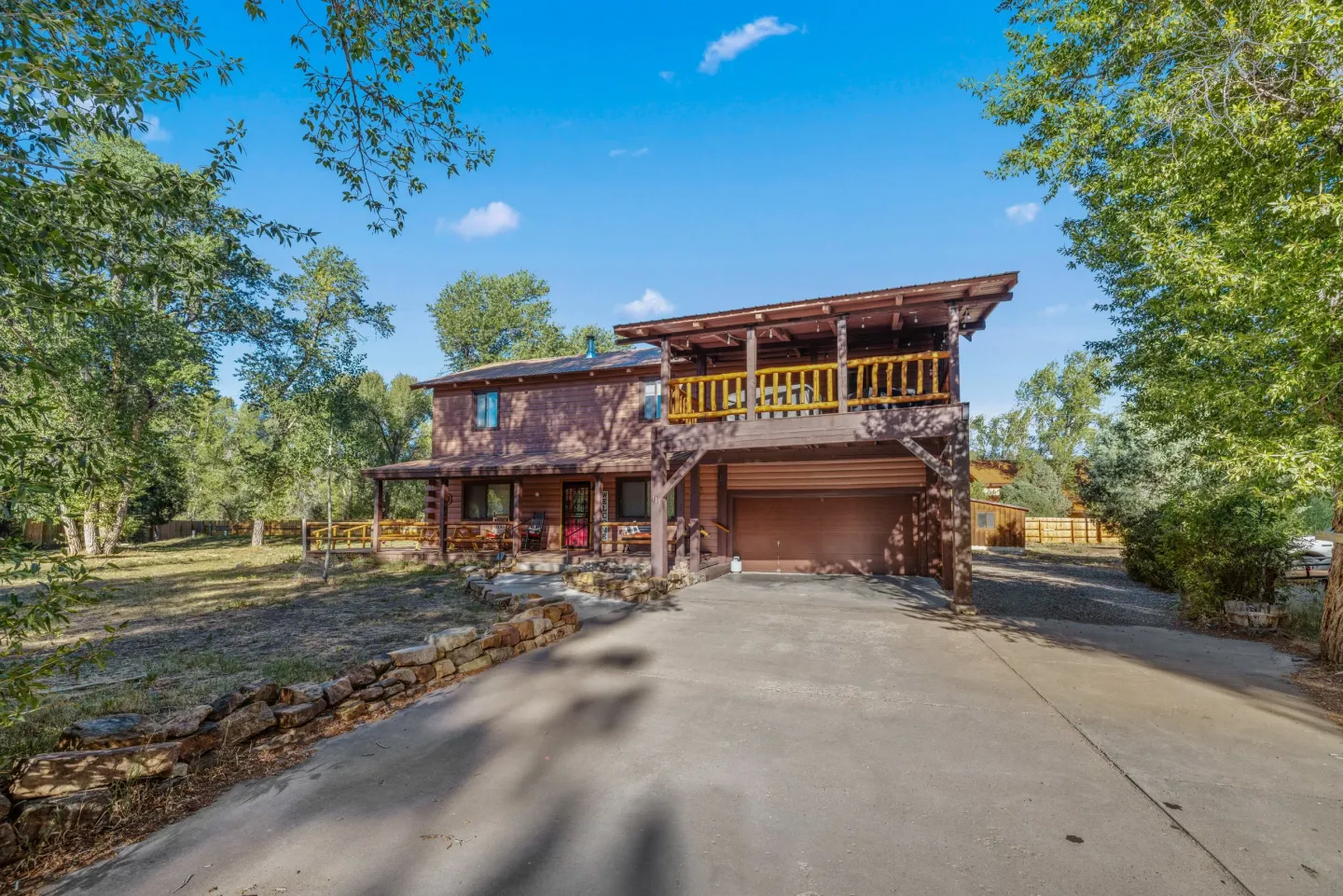 Exterior front view of a two-story wood-sided house with a garage, second-story balcony, front porch, concrete driveway, and surrounding trees under a clear blue sky.
