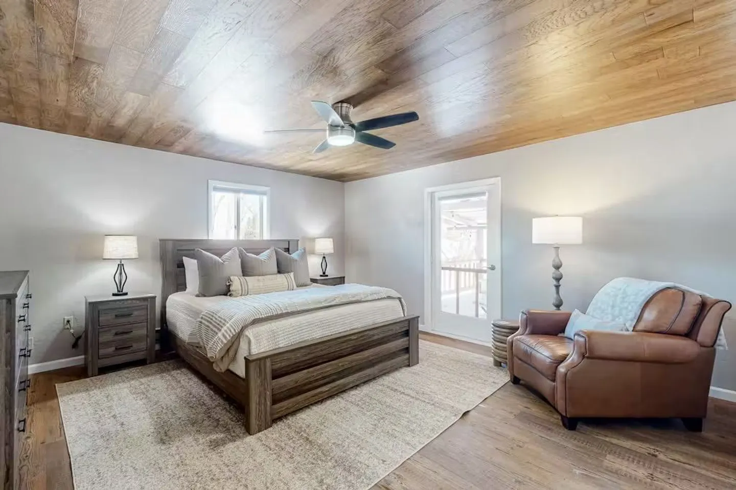 Bedroom with wood-paneled ceiling, wood-frame bed on an area rug, leather armchair, and exterior door.