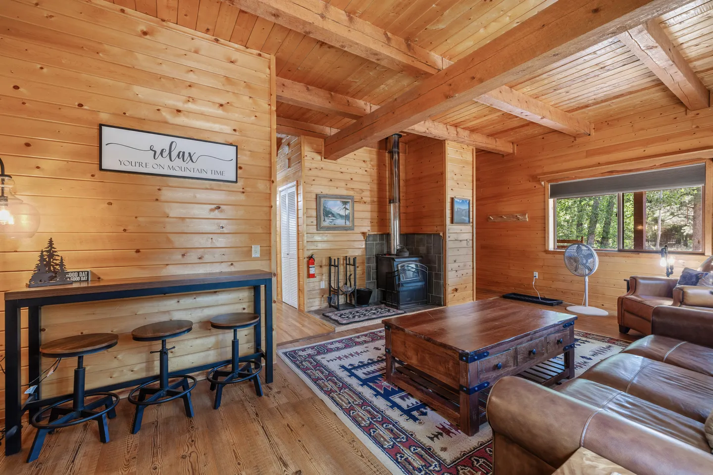 Rustic living room with wood-paneled walls and ceiling beams, leather sofa, wood-burning stove, and a high-top table with stools.