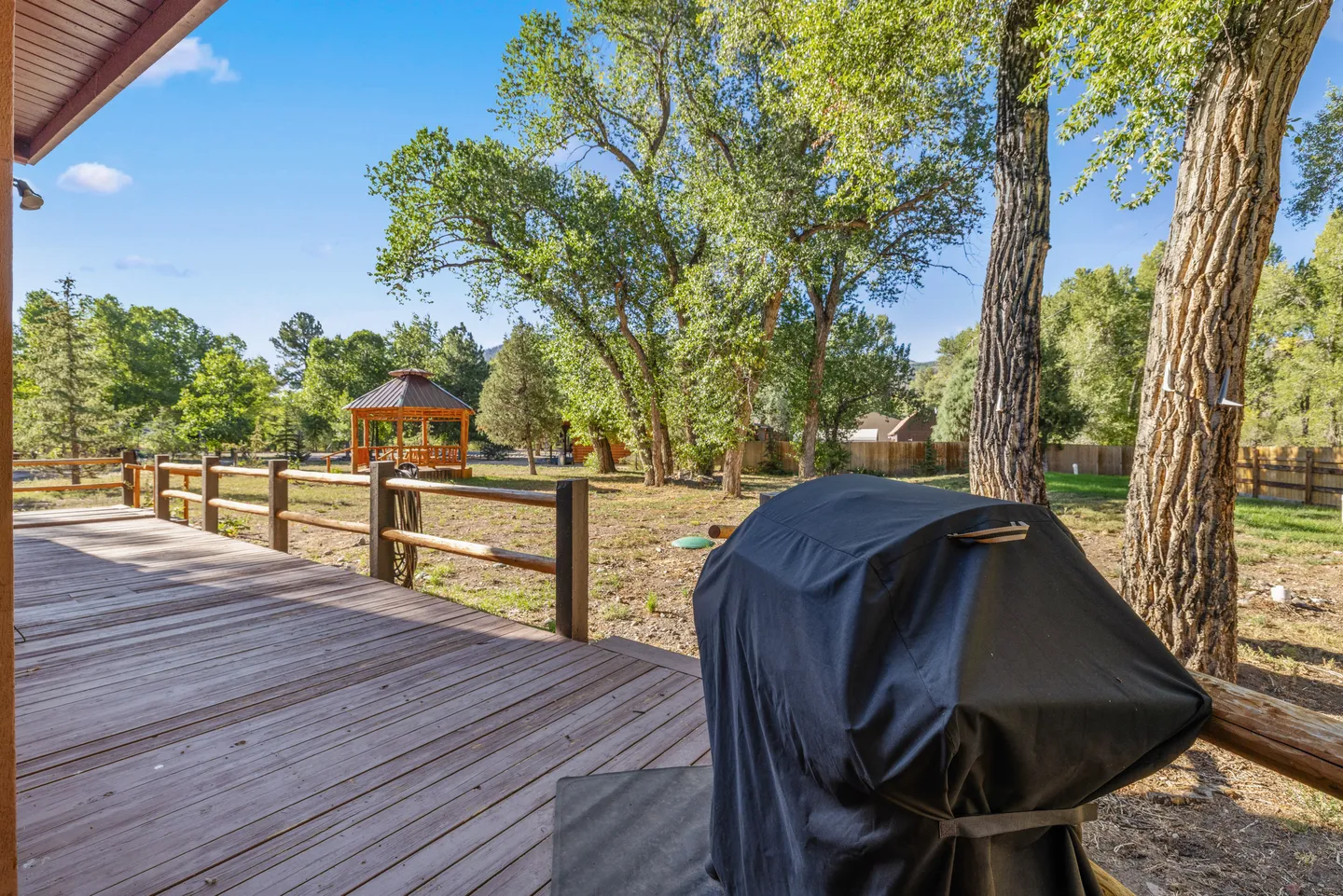 Outdoor wooden deck overlooking a fenced yard with a gazebo, mature trees, and a covered grill in the foreground.