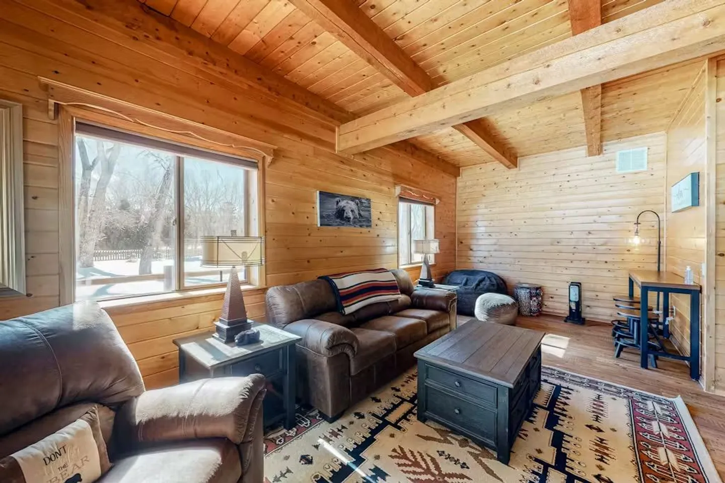 Living room with wood-paneled walls and ceiling beams, leather sofa, recliner, coffee table, and windows showing a snowy yard.