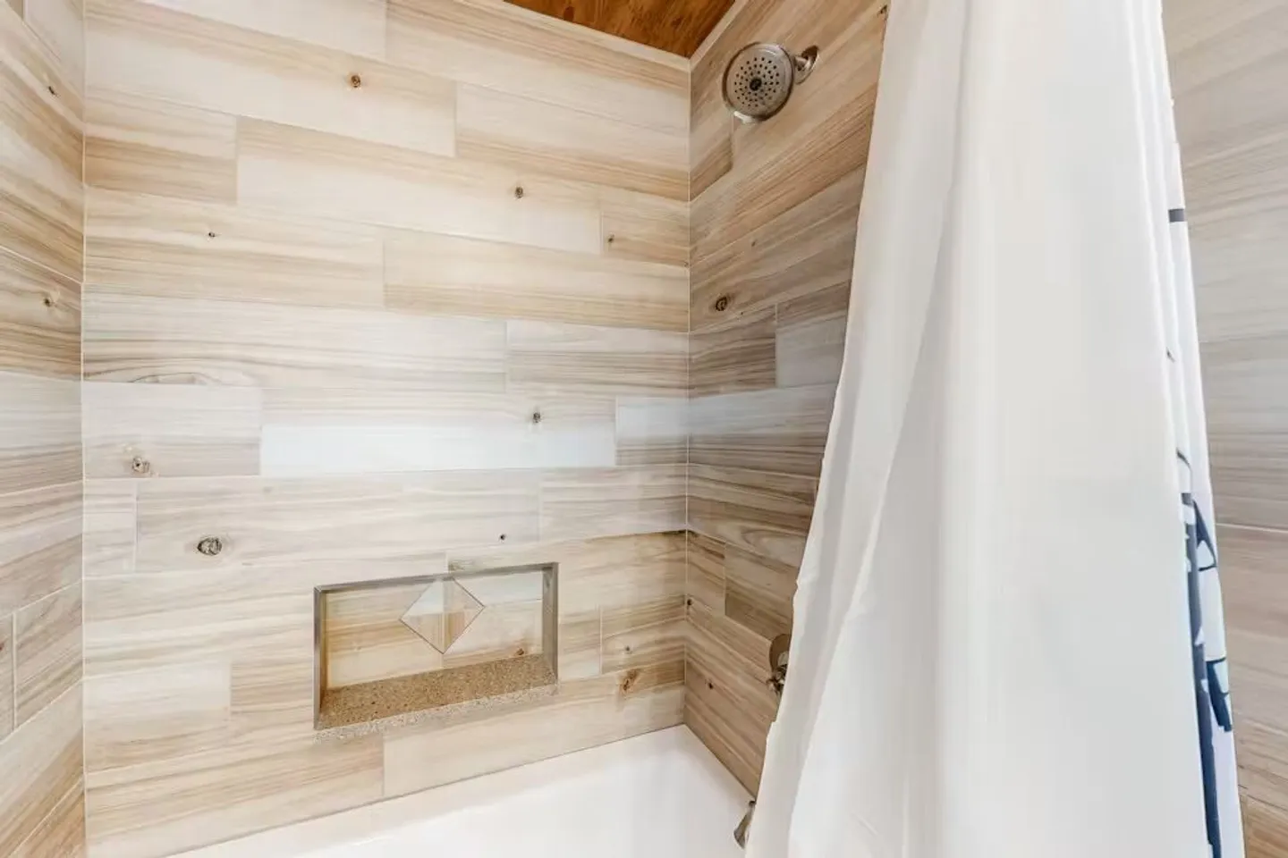 Bathroom shower and tub combination featuring wood-look tile walls, an inset shampoo niche, a wall-mounted showerhead, and a white shower curtain.