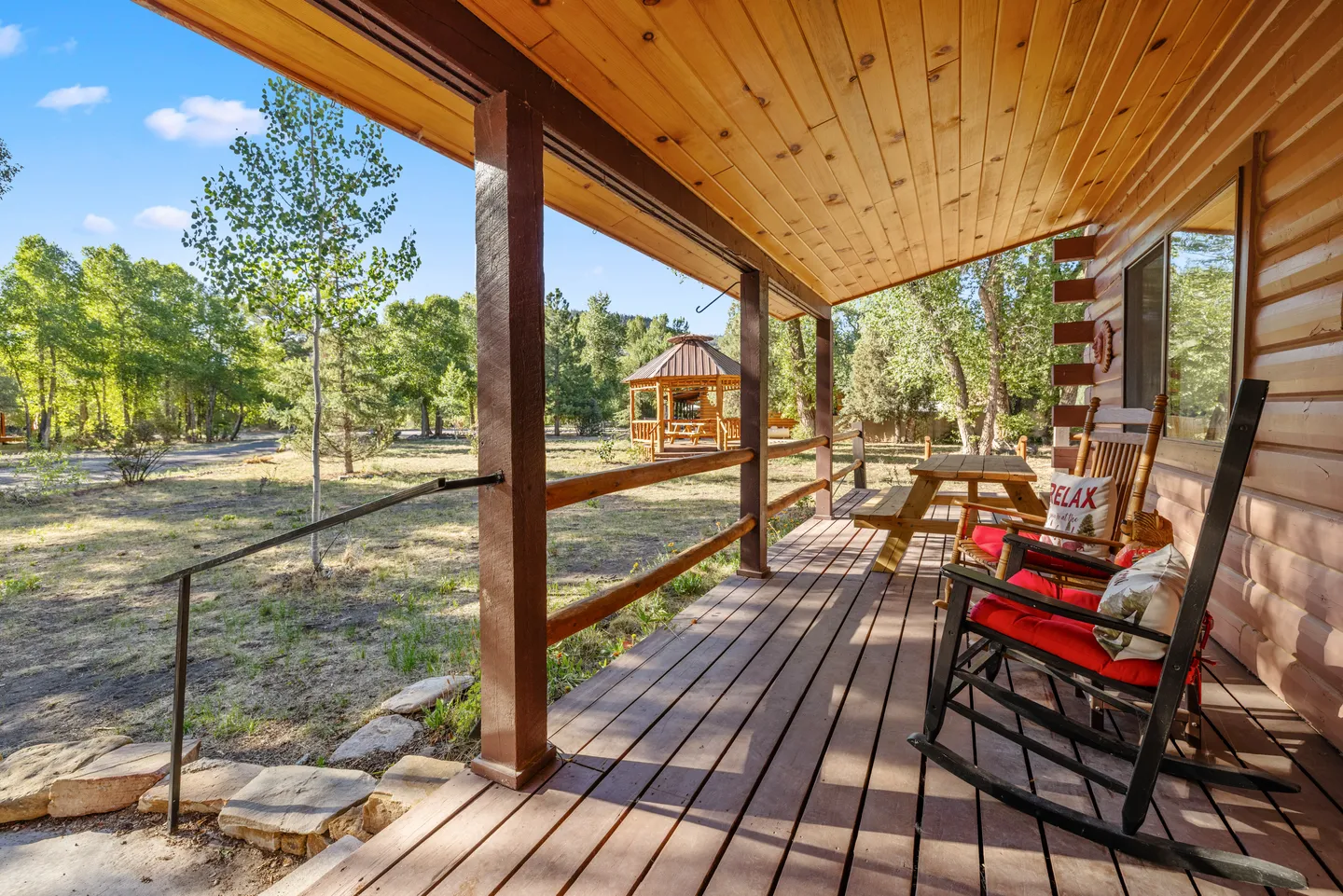Covered wooden porch with rocking chairs and a picnic table looking out toward a yard with trees and a gazebo.