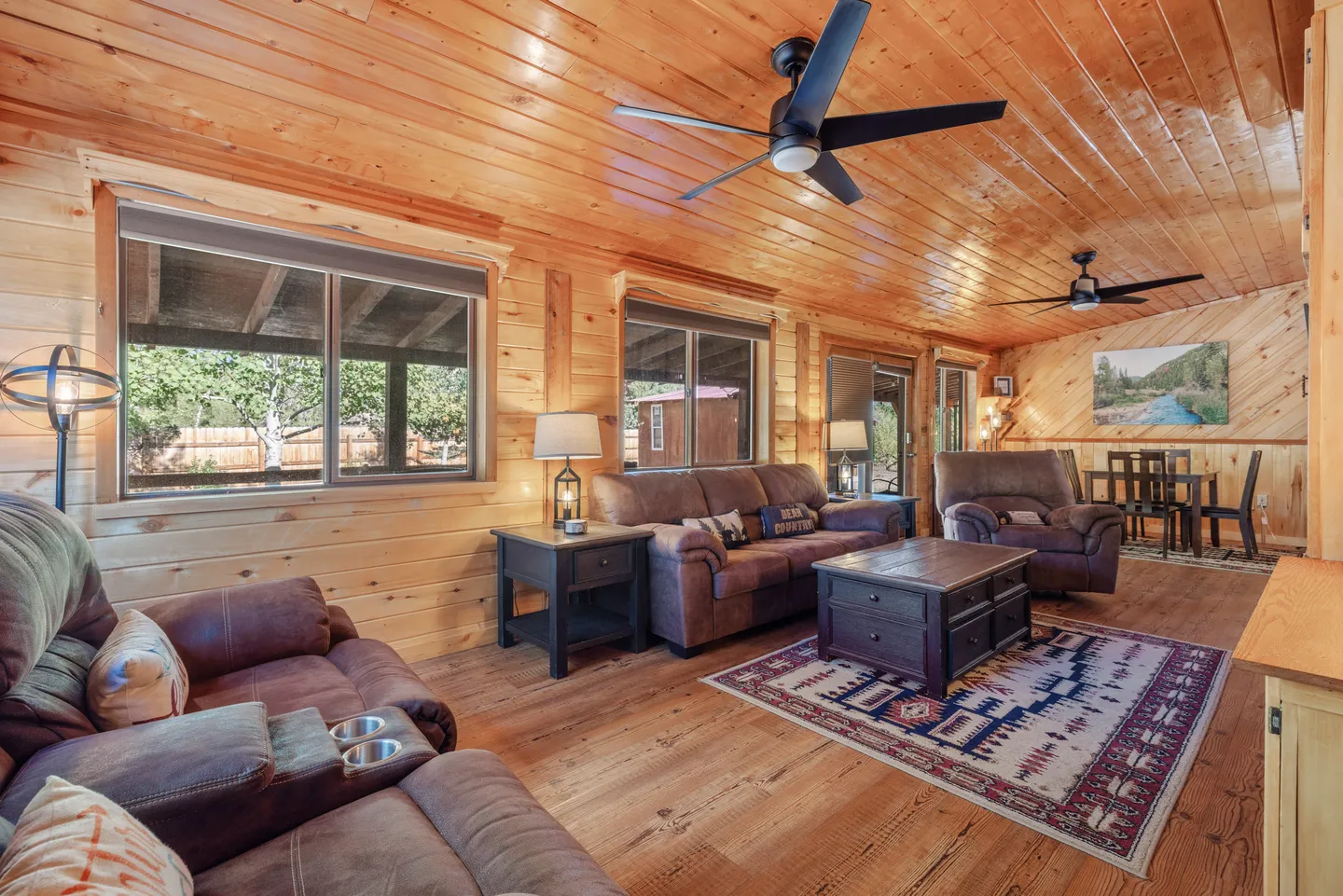Living room with knotty pine paneled walls, wood ceiling with ceiling fans, brown upholstered seating, and wood flooring.