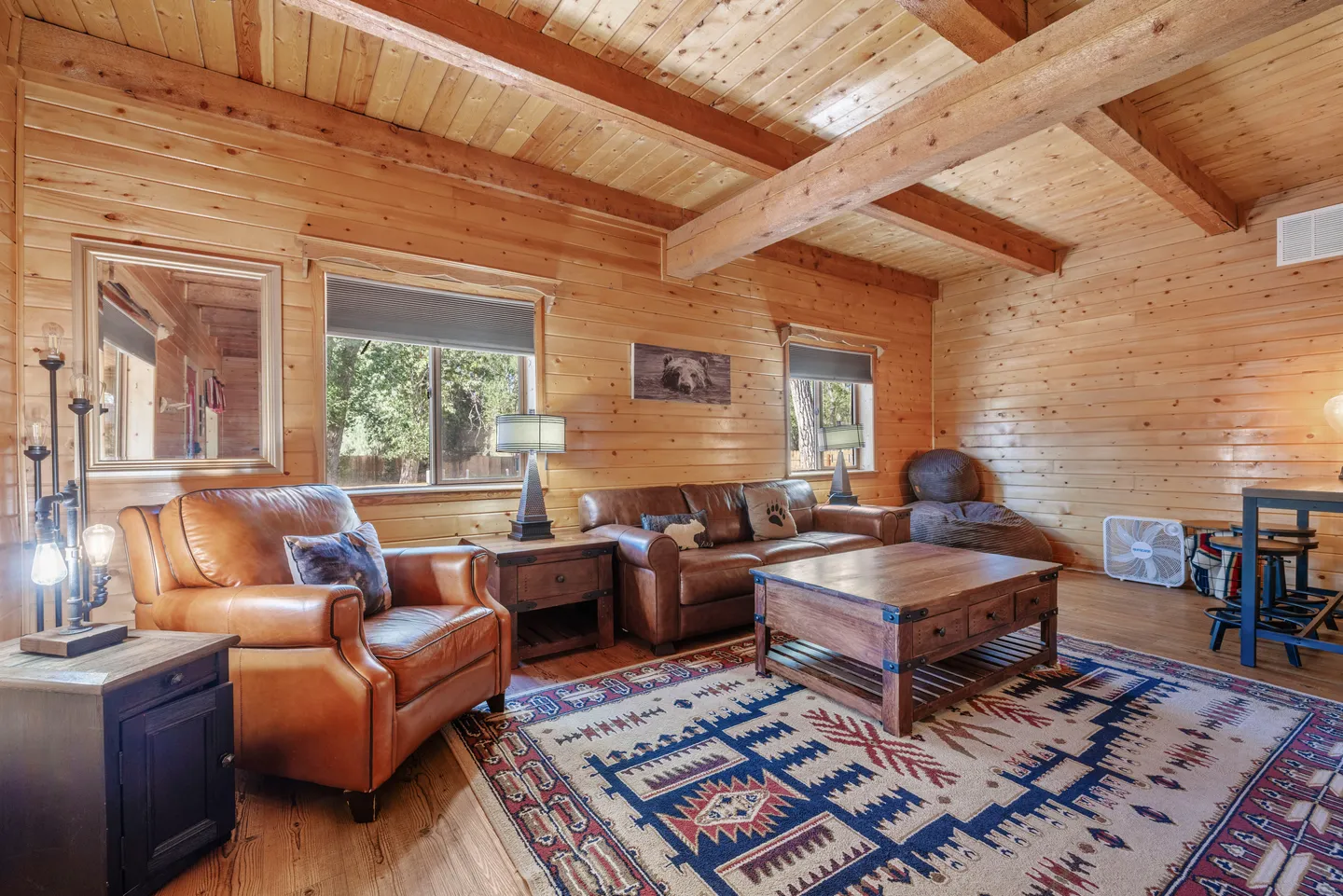 Living room with wood-paneled walls and exposed ceiling beams, furnished with leather seating, a rustic coffee table, and a patterned area rug.