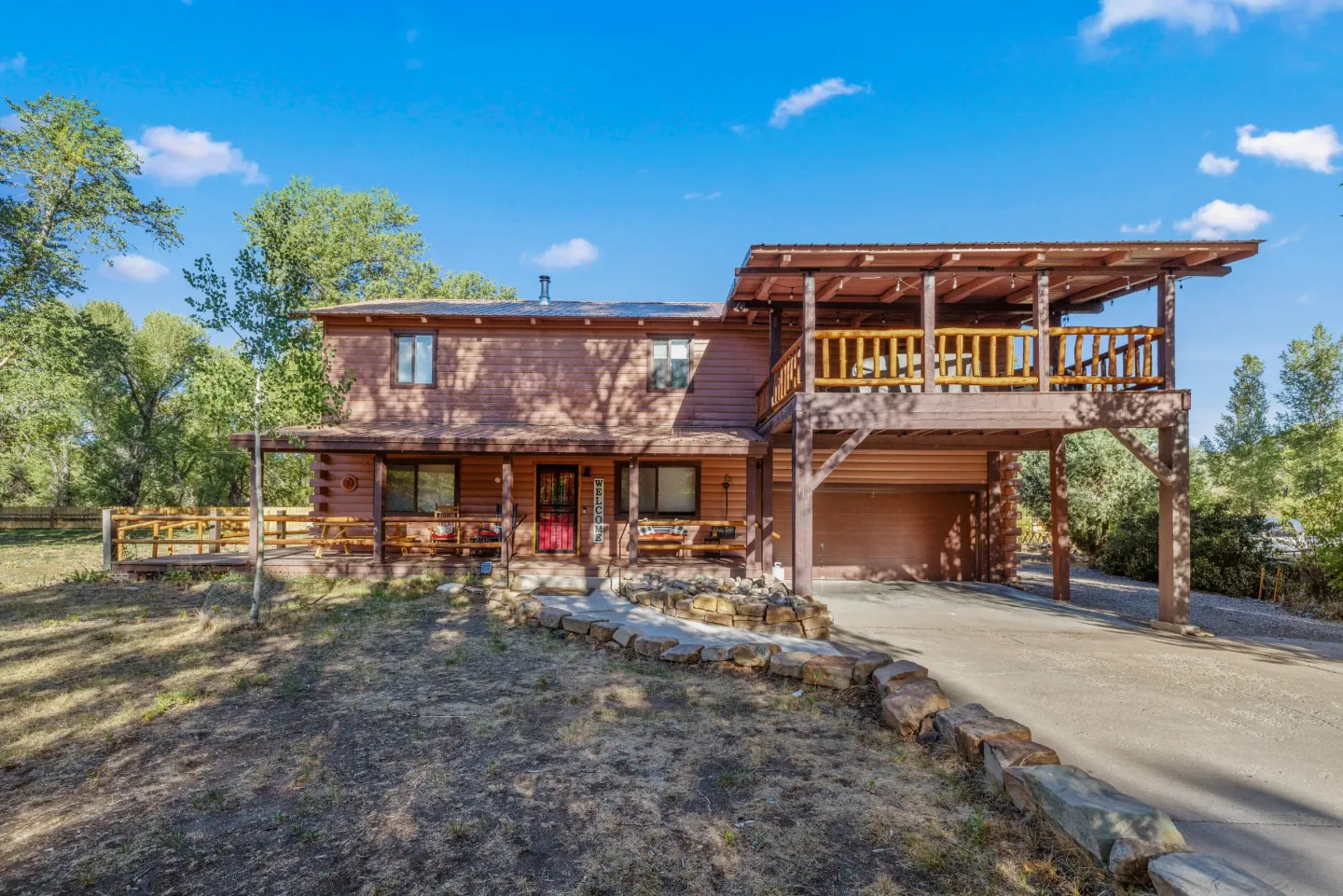 Front exterior of a two-story log-sided home with a covered front porch, an upper deck over the carport, and a stone-lined driveway.