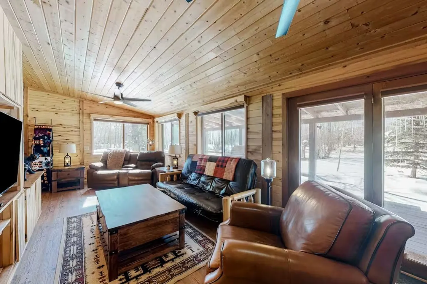 Living room with wood-paneled walls and ceiling, leather seating, a coffee table, and large windows looking outside.