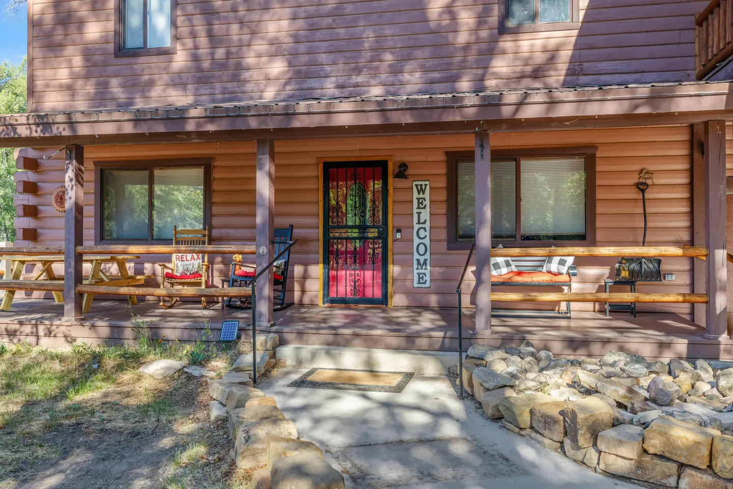 Front porch of a log cabin with outdoor seating, a red door with decorative metal screen, and a welcome sign.