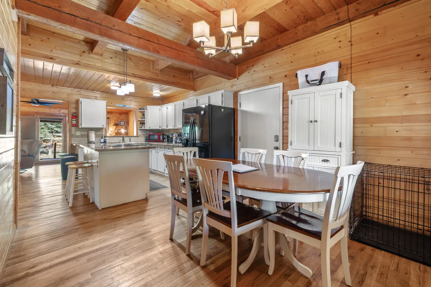 Dining area with a wooden table and white chairs leading into an open kitchen with log cabin walls and wood-beamed ceilings.