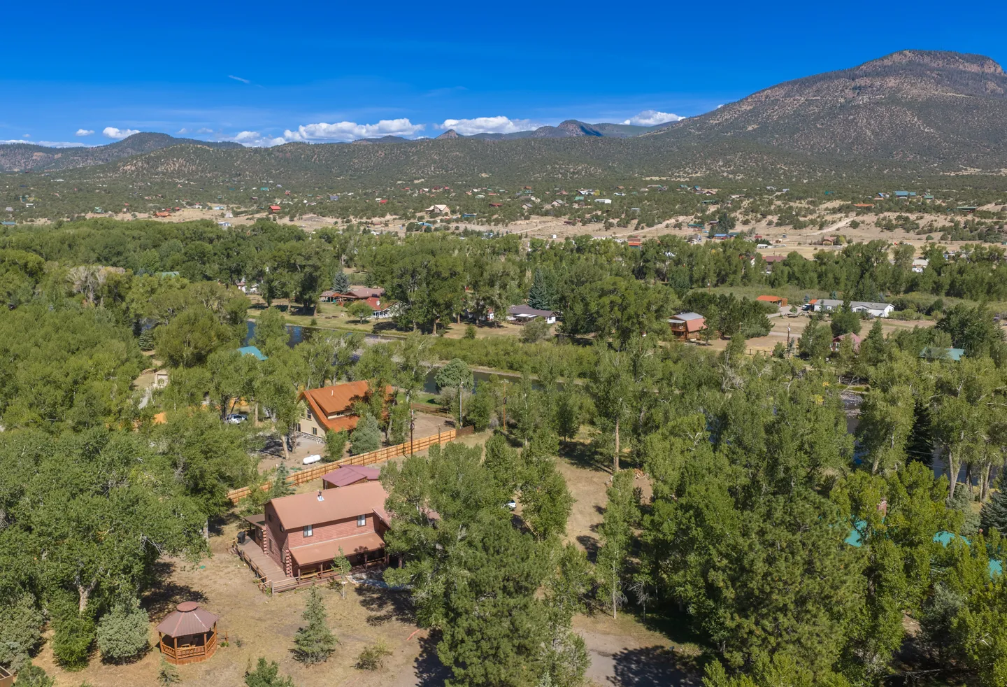 Aerial view of a cabin nestled among lush green trees with surrounding valley homes and distant mountain ranges under a blue sky.