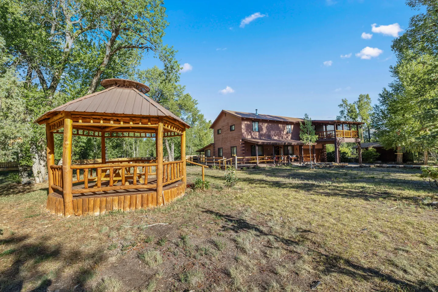 A rustic wooden gazebo in an open grassy yard with a two-story log cabin in the background under a blue sky.