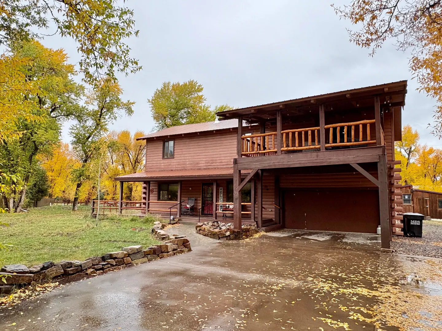 Exterior front view of a two-story log-style home with an attached garage, upper balcony, and wet asphalt driveway lined with autumn trees.