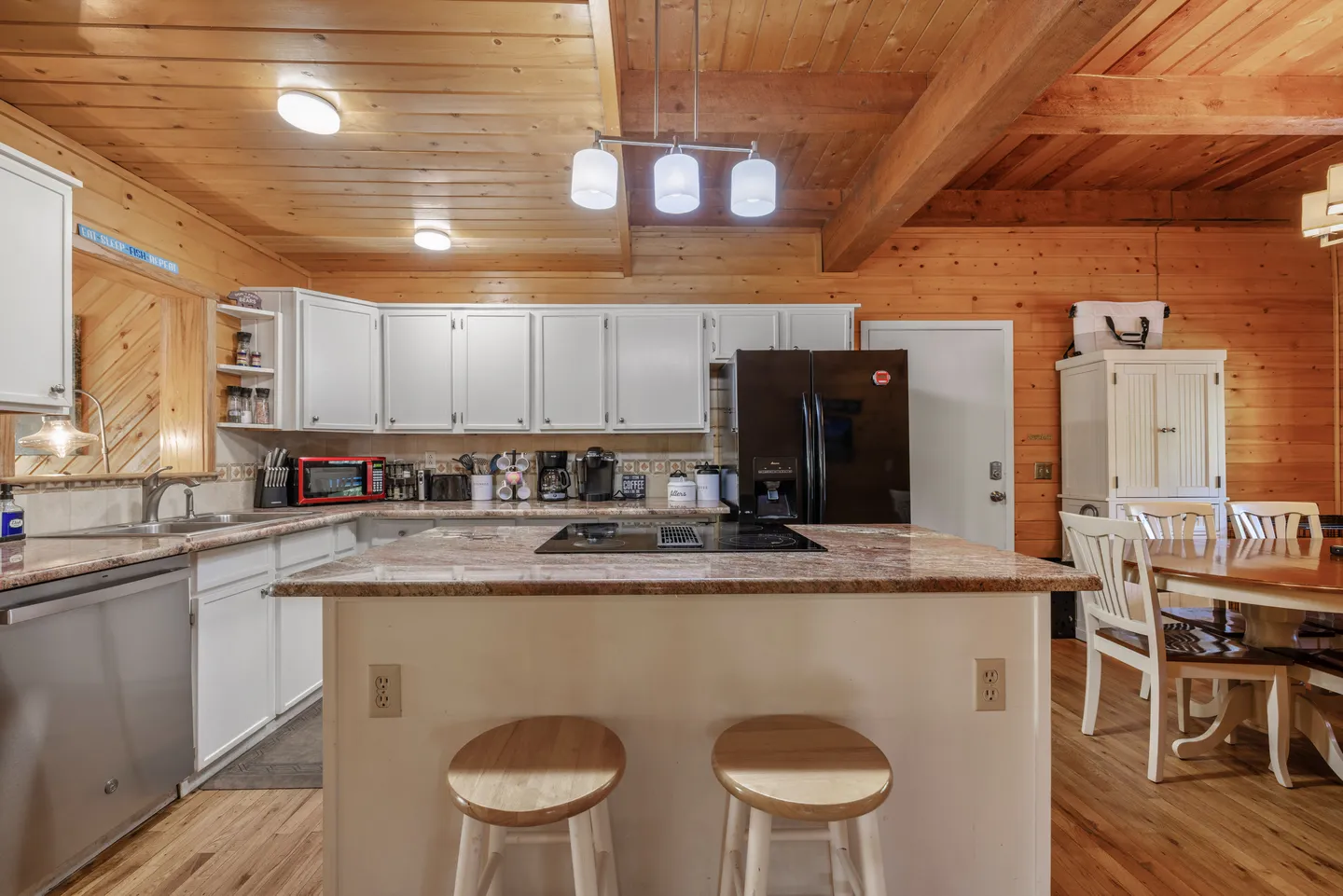 Kitchen with white cabinetry, a center island with a built-in cooktop and two round stools, wood paneled walls, and a wood plank ceiling.