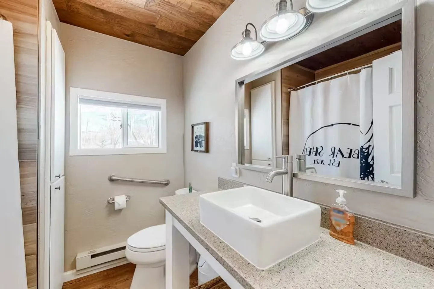 Bathroom with a rectangular vessel sink, wooden ceiling, toilet with grab bar, and small window.
