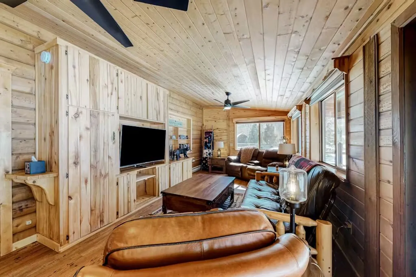 Living room with knotty wood-paneled walls and ceiling, a built-in entertainment center with a flat-screen TV, and leather seating.