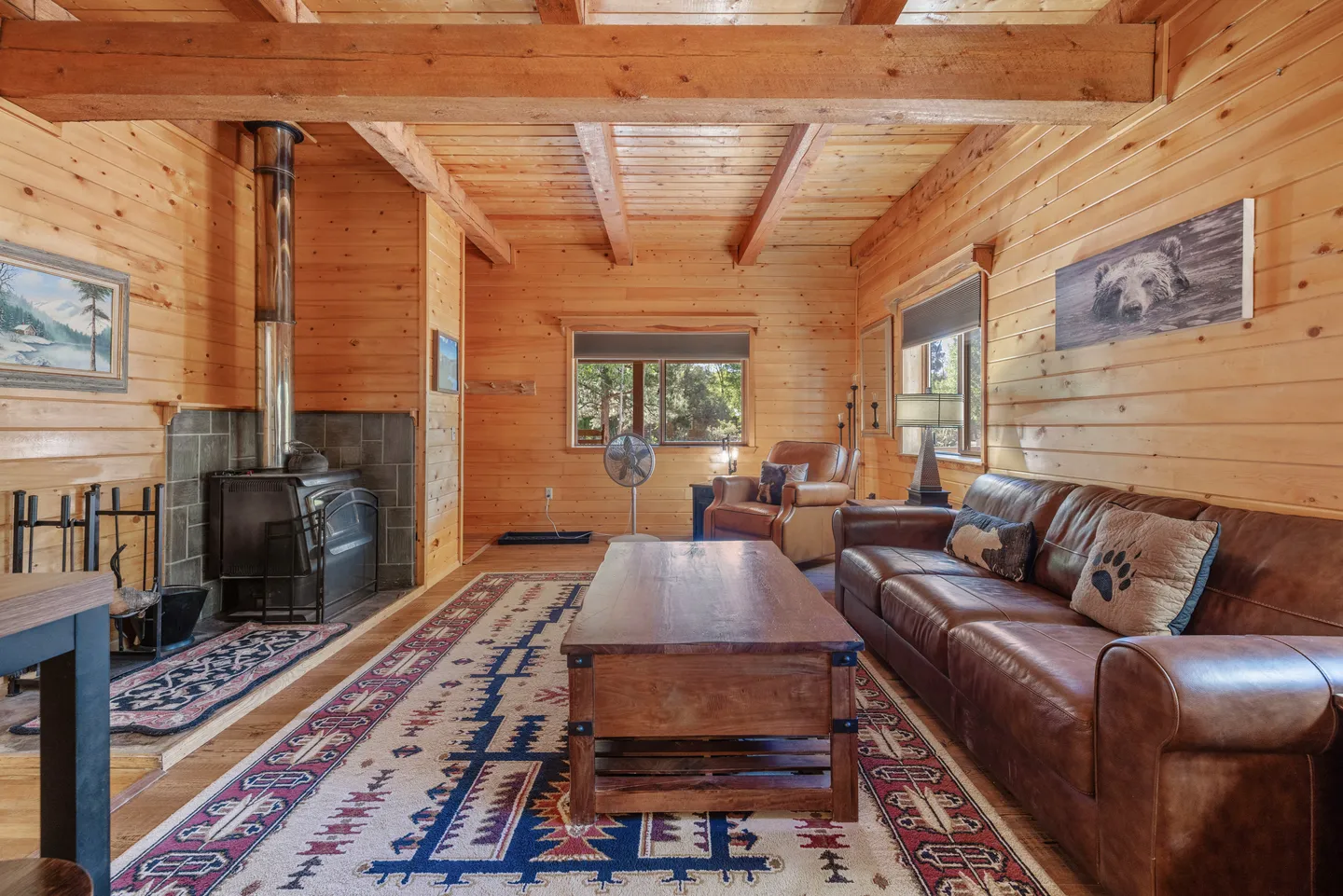 Living room with wood-paneled walls, exposed ceiling beams, a leather couch, a wood stove, and a patterned rug.