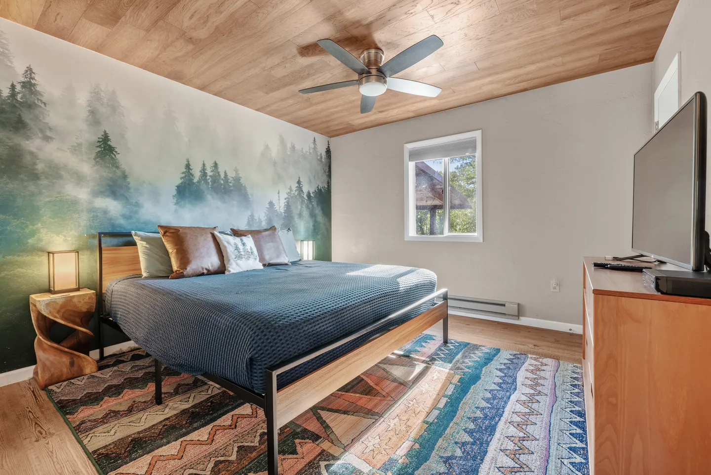 Bedroom featuring a bed with a blue textured spread, a forest-themed wall mural, wood-paneled ceiling with a fan, and a flat-screen TV.
