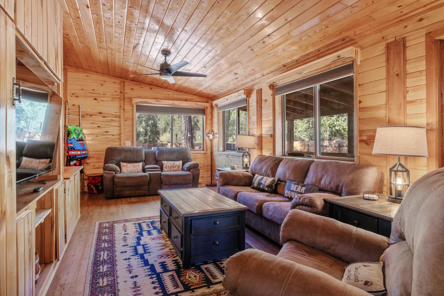 Living room with wood-paneled walls and ceiling, brown reclining sofas, and a wooden coffee table over an area rug.