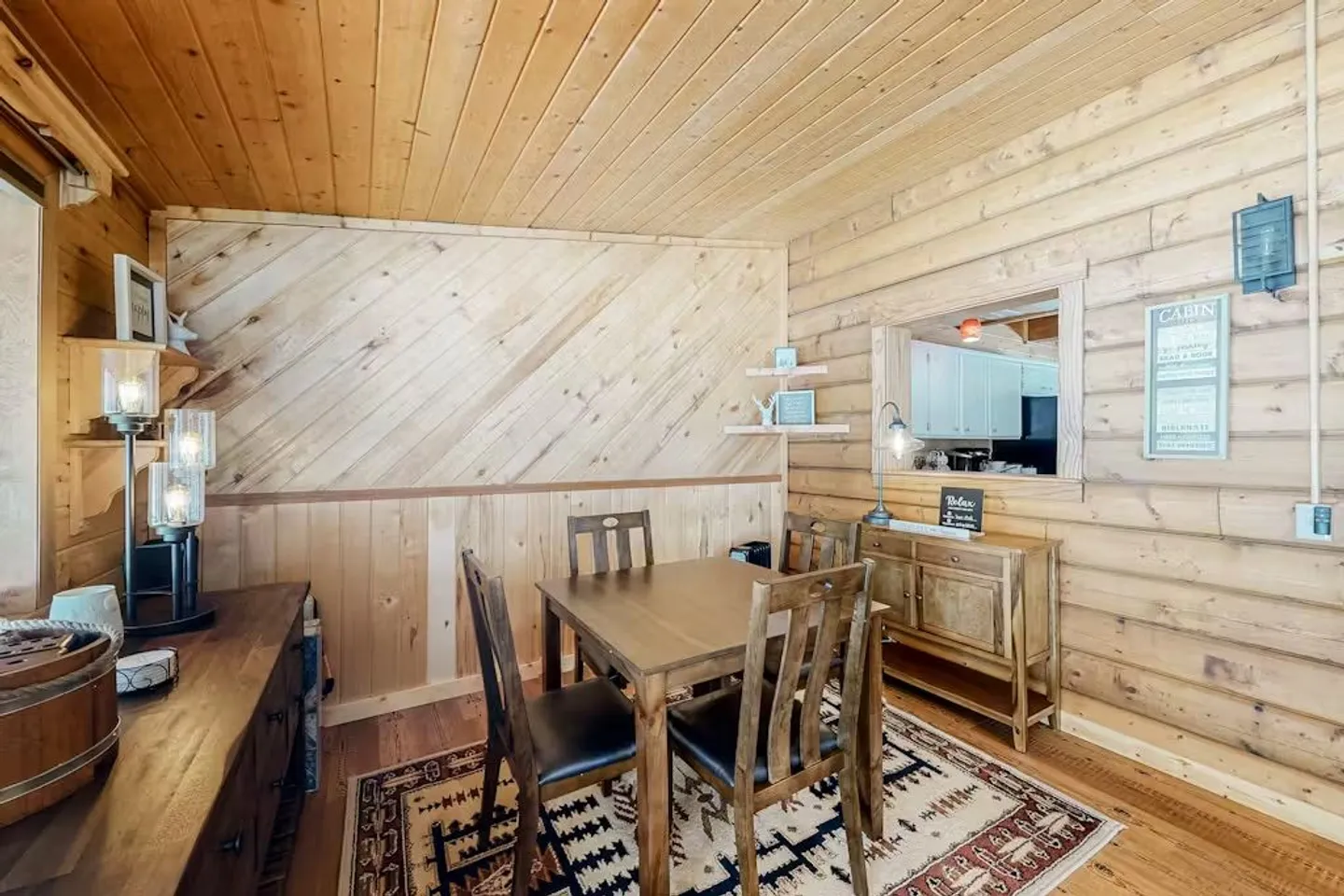 Dining area with wood-paneled walls and ceiling, a square wooden dining table, chairs, a patterned rug, and a pass-through window to the kitchen.