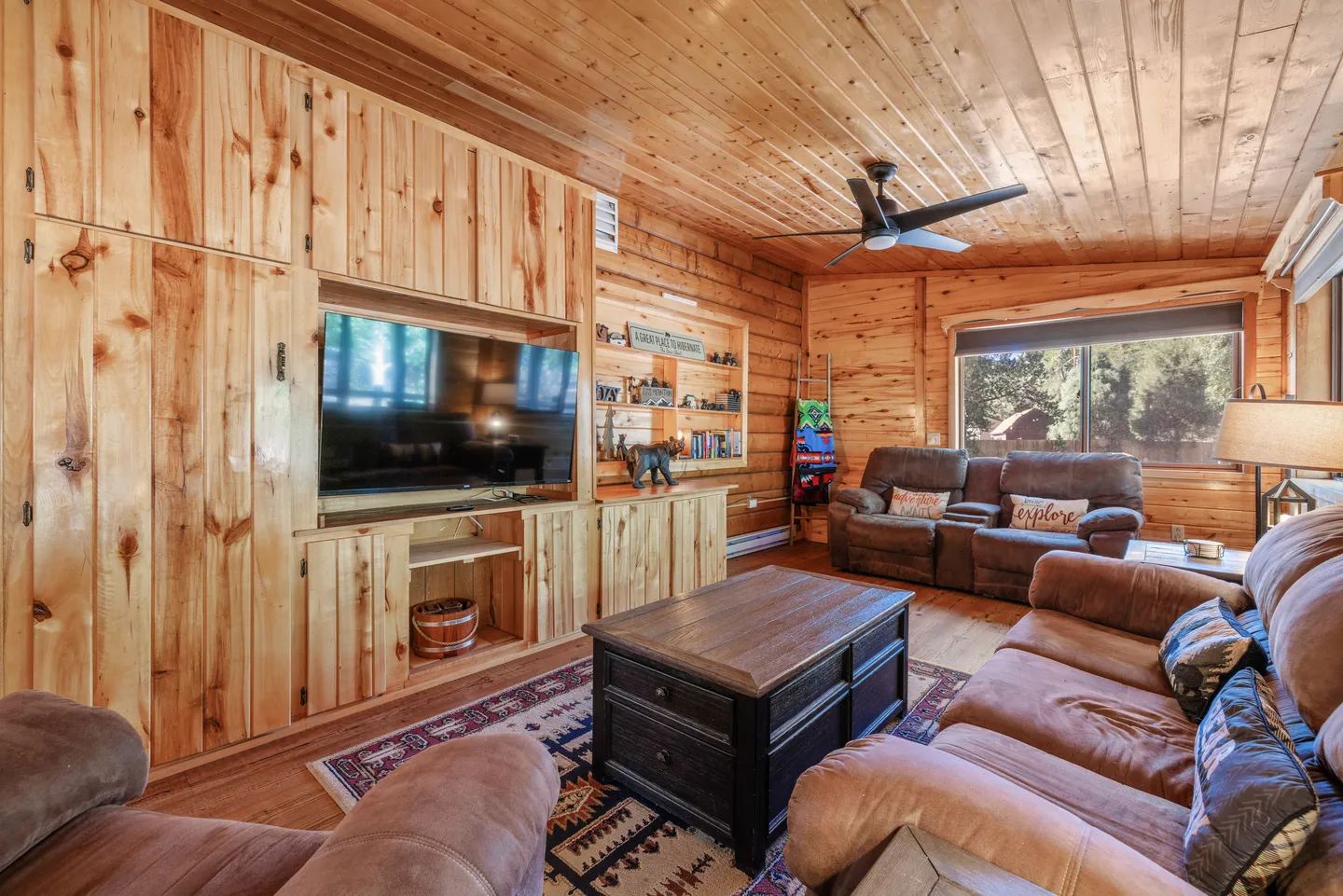 Living room with knotty pine wood-paneled walls and ceiling, brown reclining sofas, a coffee table, and a built-in entertainment center with a flat-screen TV.