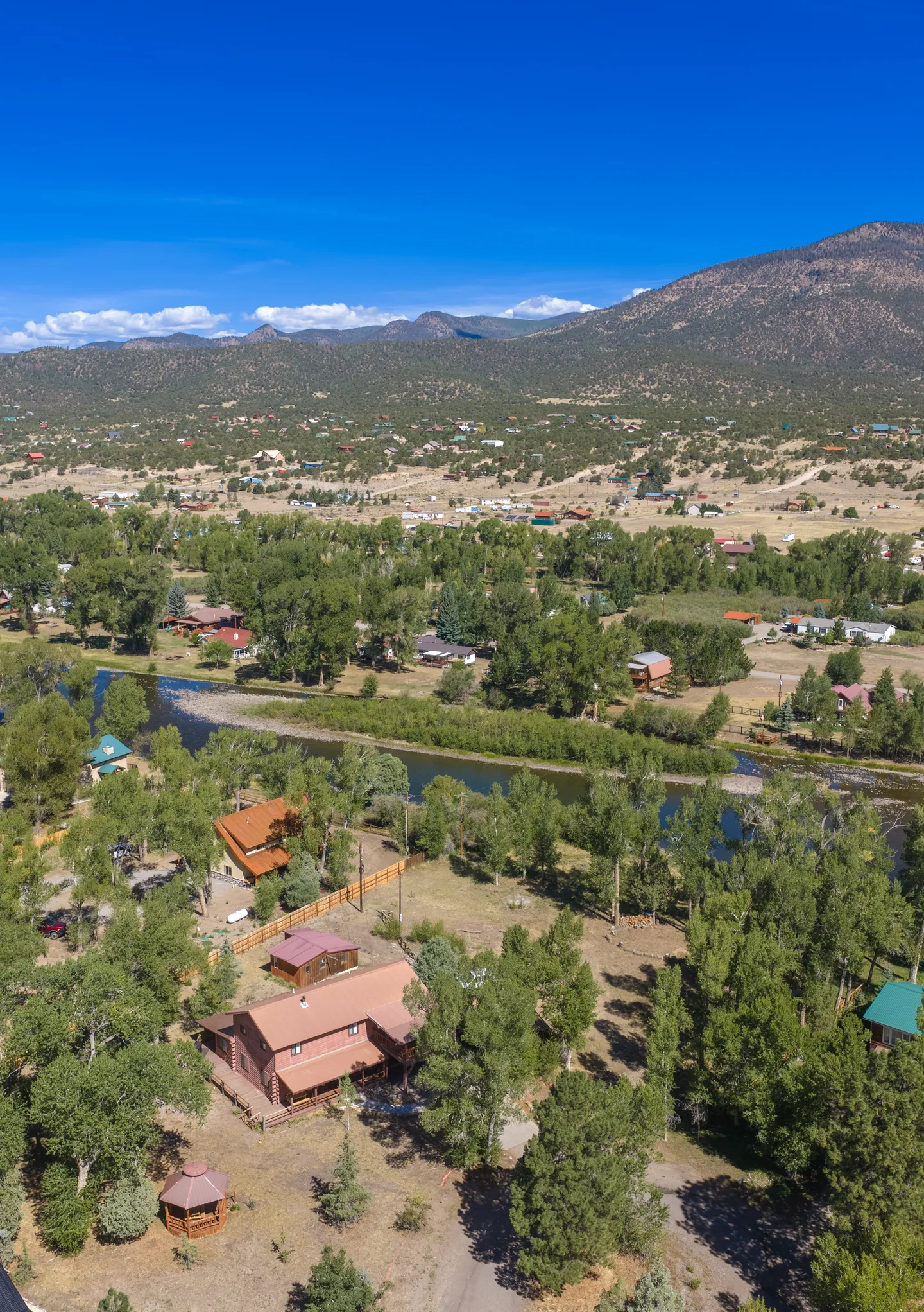 Aerial view of a log cabin neighborhood along a river with mountains in the background under a blue sky.