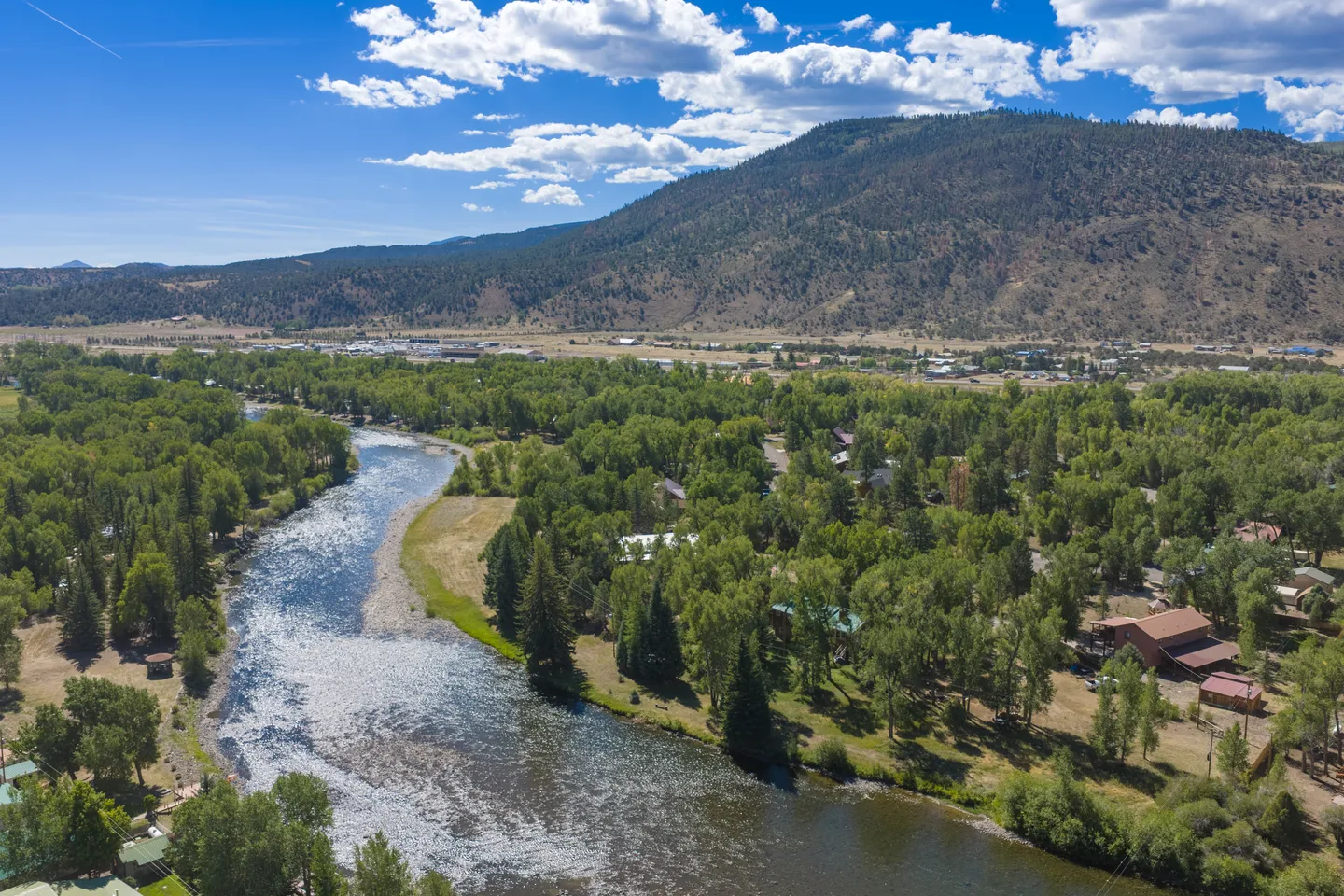 Aerial view of a wide river curving through a lush green valley with trees, houses, and mountains in the background under a blue sky.