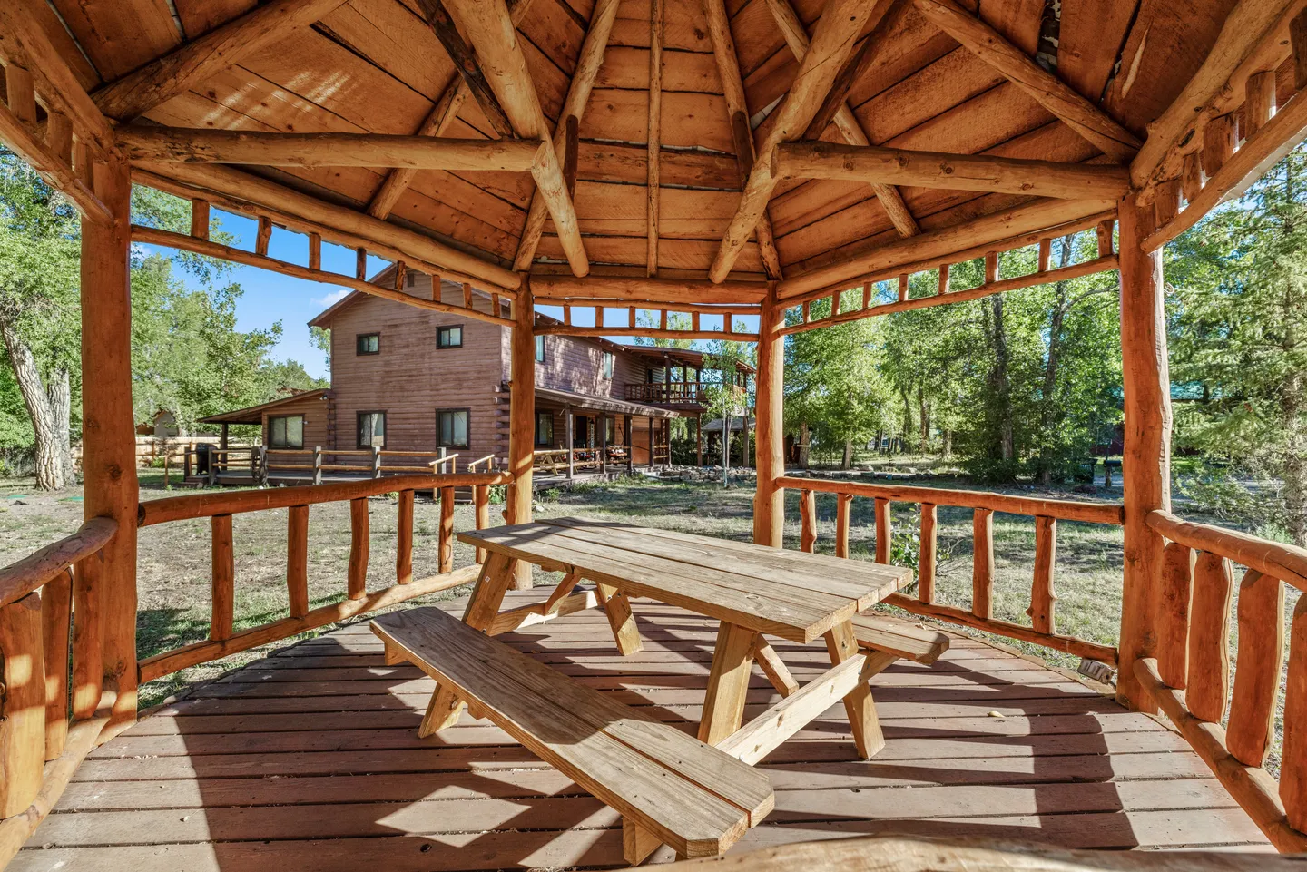 Interior view of a rustic log gazebo with a wooden picnic table, looking out toward a two-story log house.