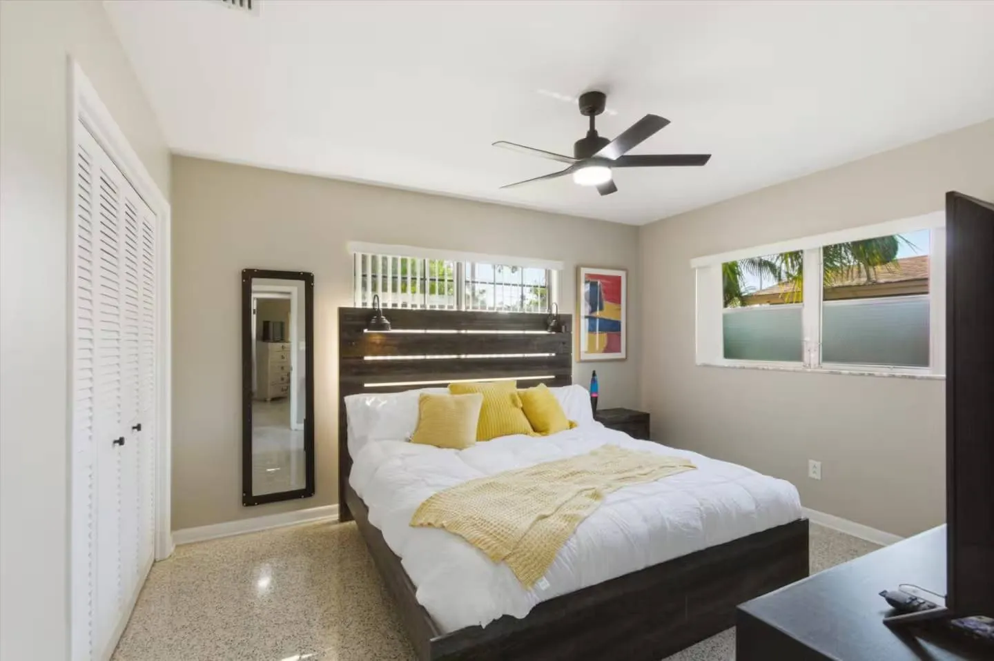 Bedroom with a dark wood bed frame, white bedding with yellow pillows, ceiling fan, and louvered closet doors.