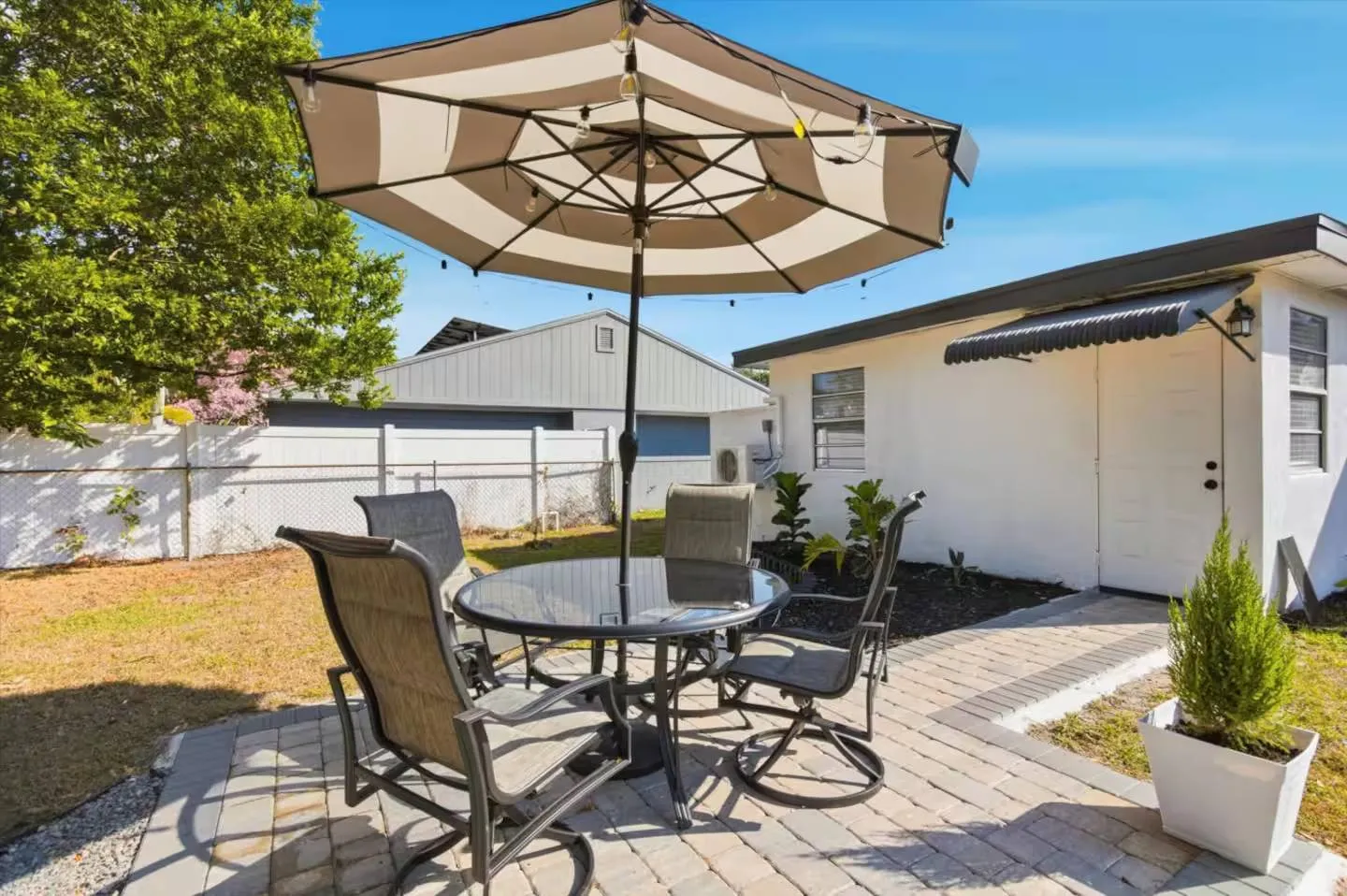 Outdoor patio with a paved stone ground, round glass-top table, chairs, and a large umbrella in a fenced backyard.