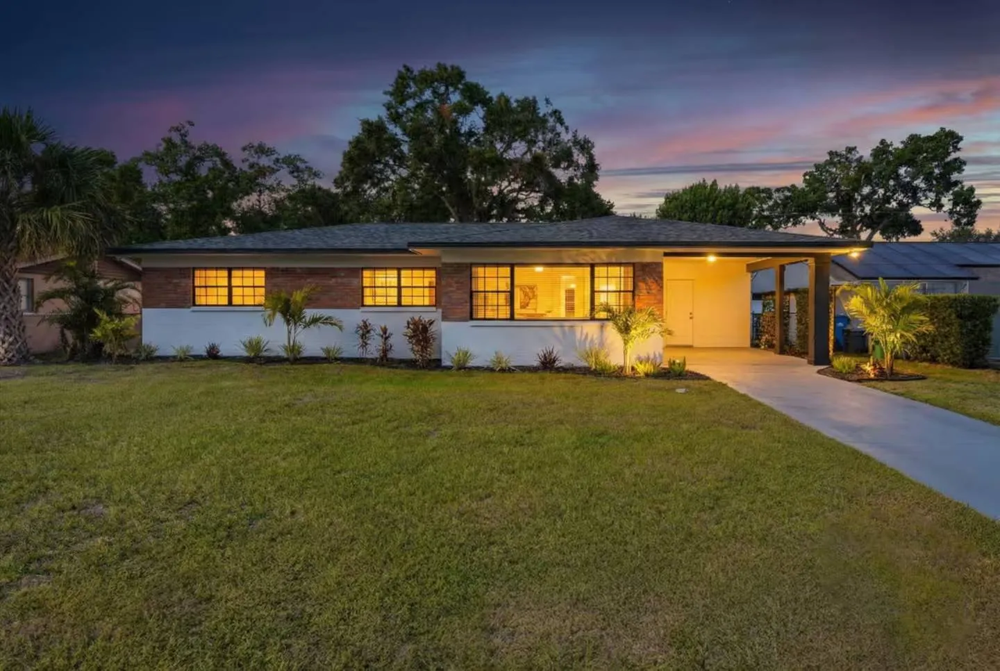 Exterior front view of a single-story home at twilight with lit windows, a carport, and a green lawn.