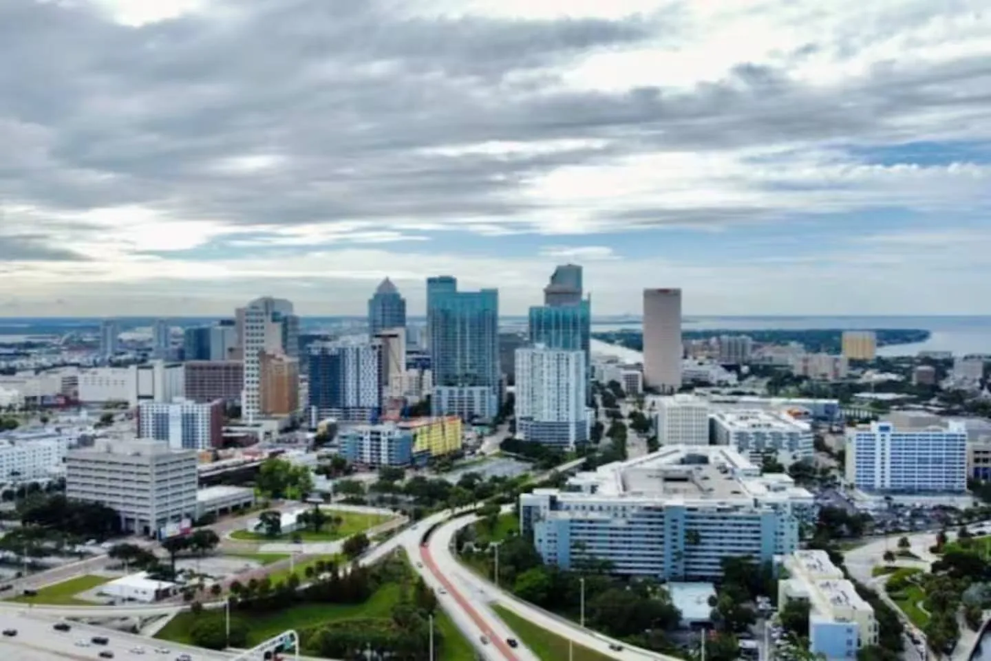 Aerial view of a city skyline with skyscrapers, highways, and water in the background under a cloudy sky.