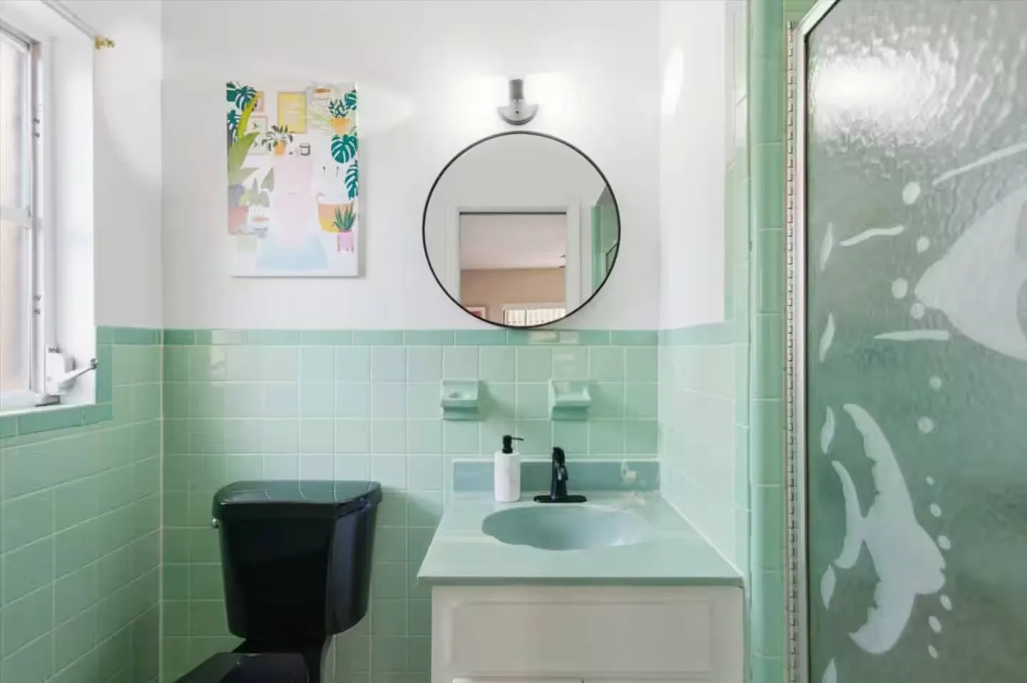 Bathroom with mint green wall tiles, white vanity, round mirror, and a black toilet.
