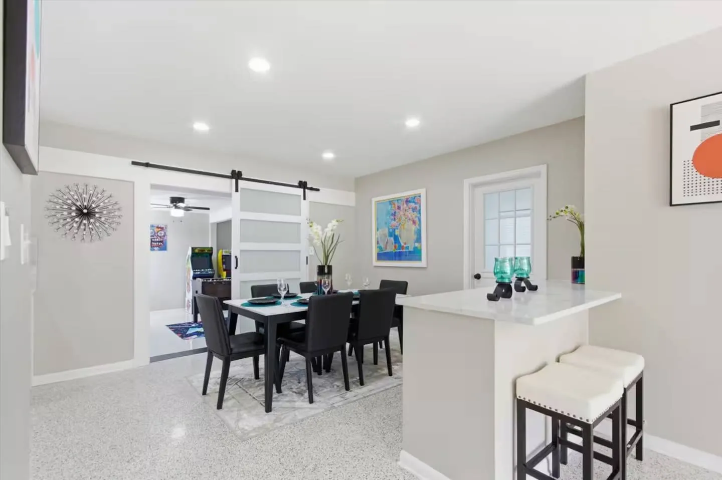 Dining area with a black table and chairs, adjacent breakfast bar with stools, and sliding barn door leading to a game room.