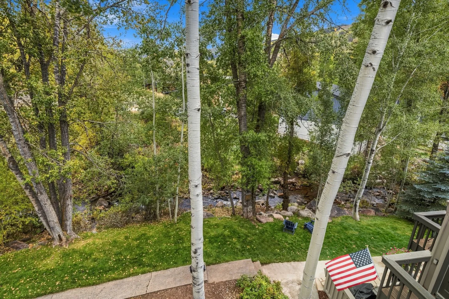 Elevated outdoor view through birch trees overlooking a green lawn, patio area, and a rocky creek.