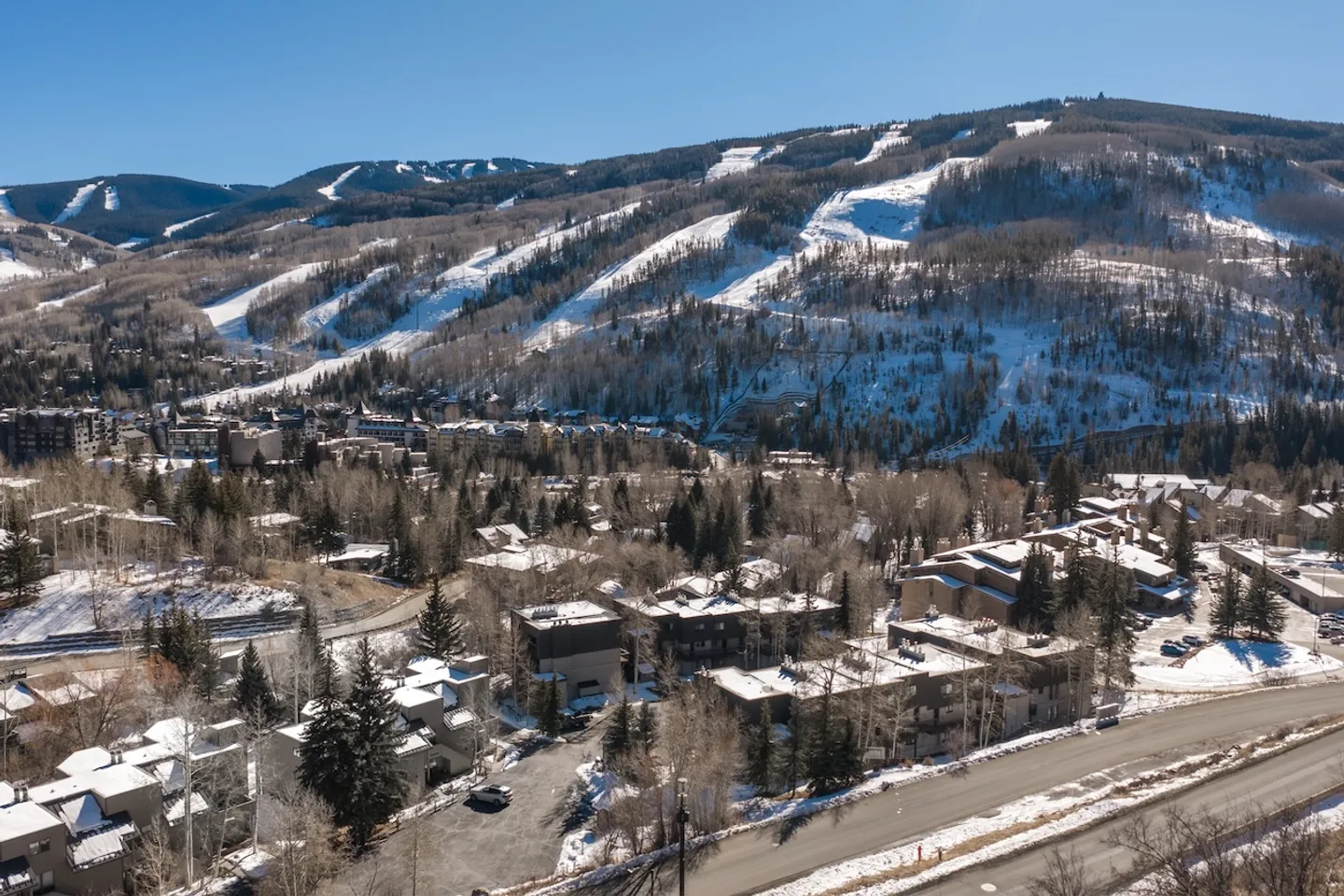 Aerial view of a mountain resort town and residences with snow-covered ski slopes in the background under a clear blue sky.